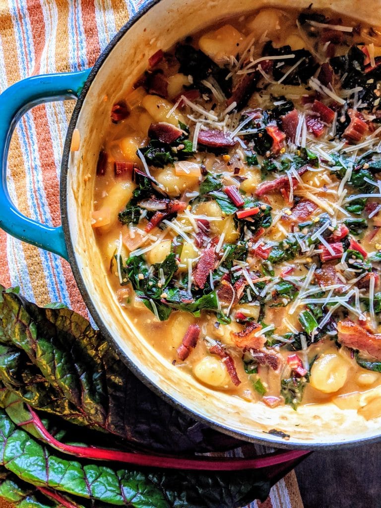 Gnocchi and swiss chard with bacon in parmesan broth in pan with striped cloth and swiss chard leaves in lower corner